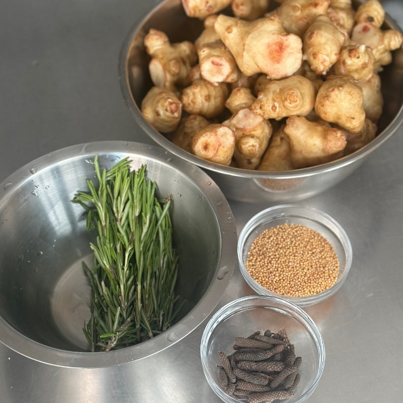 Bowls of various ingredients including sunchoke, rosemary, and mustard seeds and long pepper on a metallic surface.